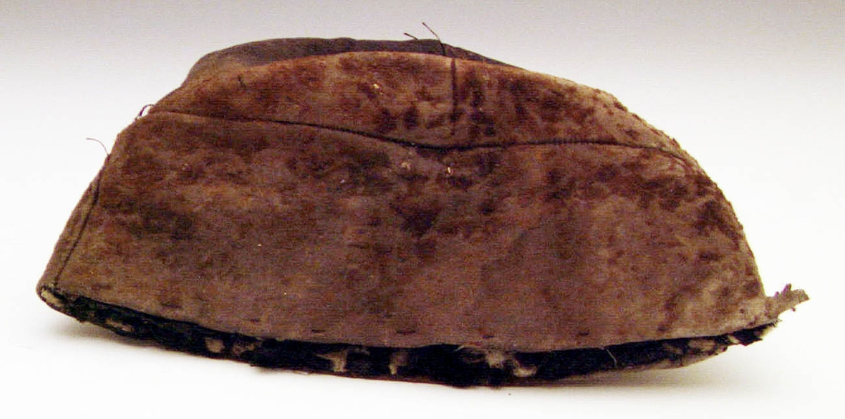 Mansmössa av skinn med rester av brun hårbeklädnad. Mössan är sydd av kilar (med många inkilade skarvar) och fodrad med vadderat, svart bomullstyg fastsytt på pappstomme. Mössan är tillplattad och något trasig. Den ger ett ålderdomligt intryck - tidigt 1800-tal.