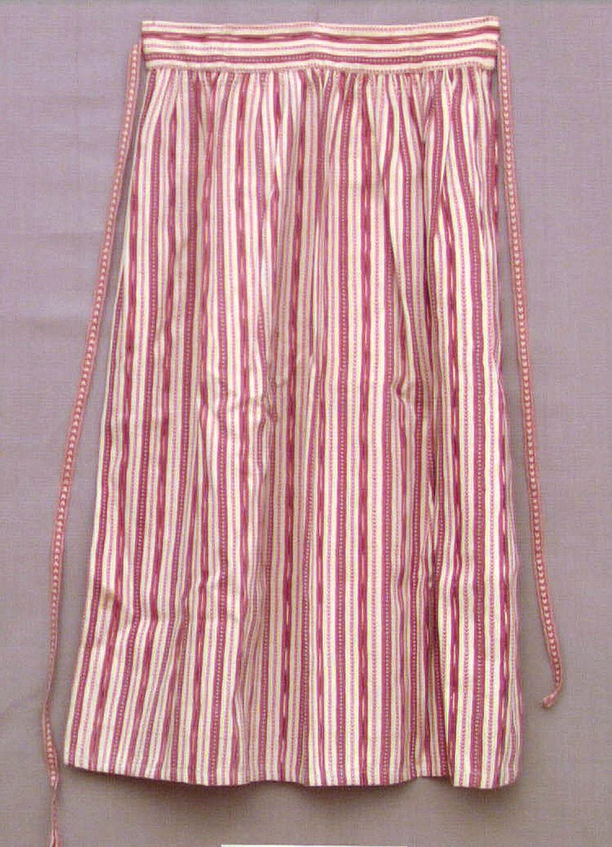 Förkläde av bomullstyg. Vit botten i tuskaft med inslag av smala ränder i rött och blått. Handvävt tyg. Försett med linning mot vilken förklädet är rynkat. Försett med handvävda bomullsband, s k "hjärtband". Förklädet är både maskin- och handsytt. Förmodligen tillverkat i modern tid. 
Ett s k "kvistaförkläde" från Ovansjö socken, använt som sommarförkläde för ogift kvinna i kyrkan.