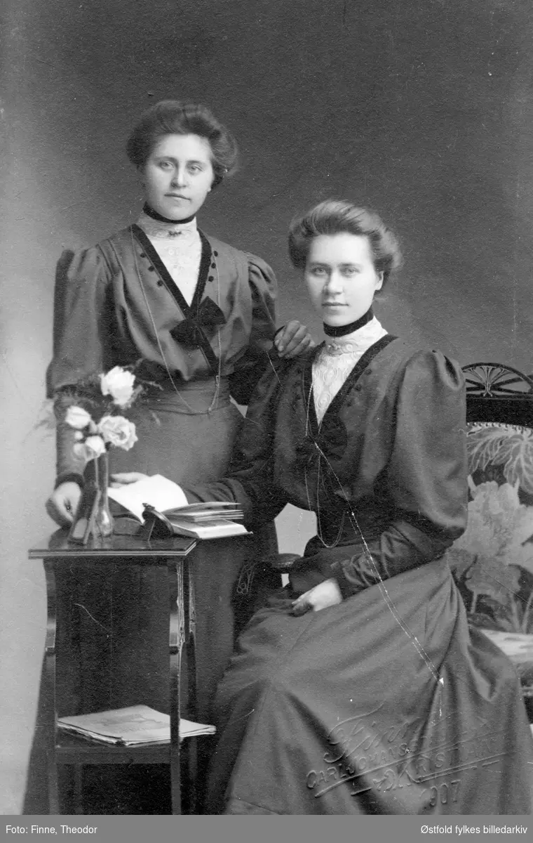 Portrett i atelier av to ukjente kvinner, visittkort, iscenesatt bilde med bord utstilt med bøker, etter 1907.