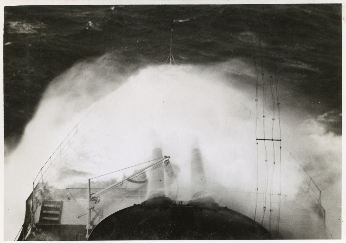 Fotot visar en fartygsför där en vattenkaskad täcker däcket. I bild syns också en del av kanonerna på däck. Bilden är tagen uppifrån en högre punkt på fartyget.