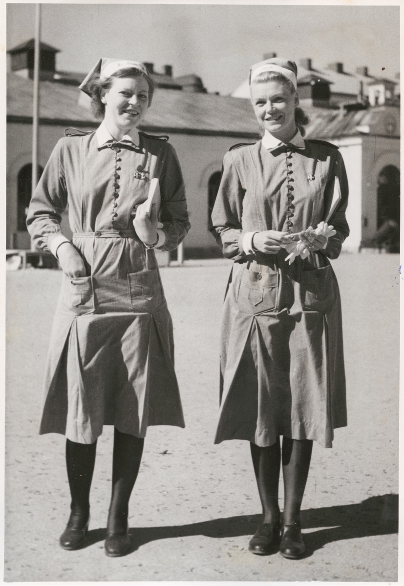Två uniformsklädda lottor fotograferade på bataljon Sparres kaserngård. Båda ler in i kameran. I bakgrunden syns exercishuset tillhörande bataljon Sparre.