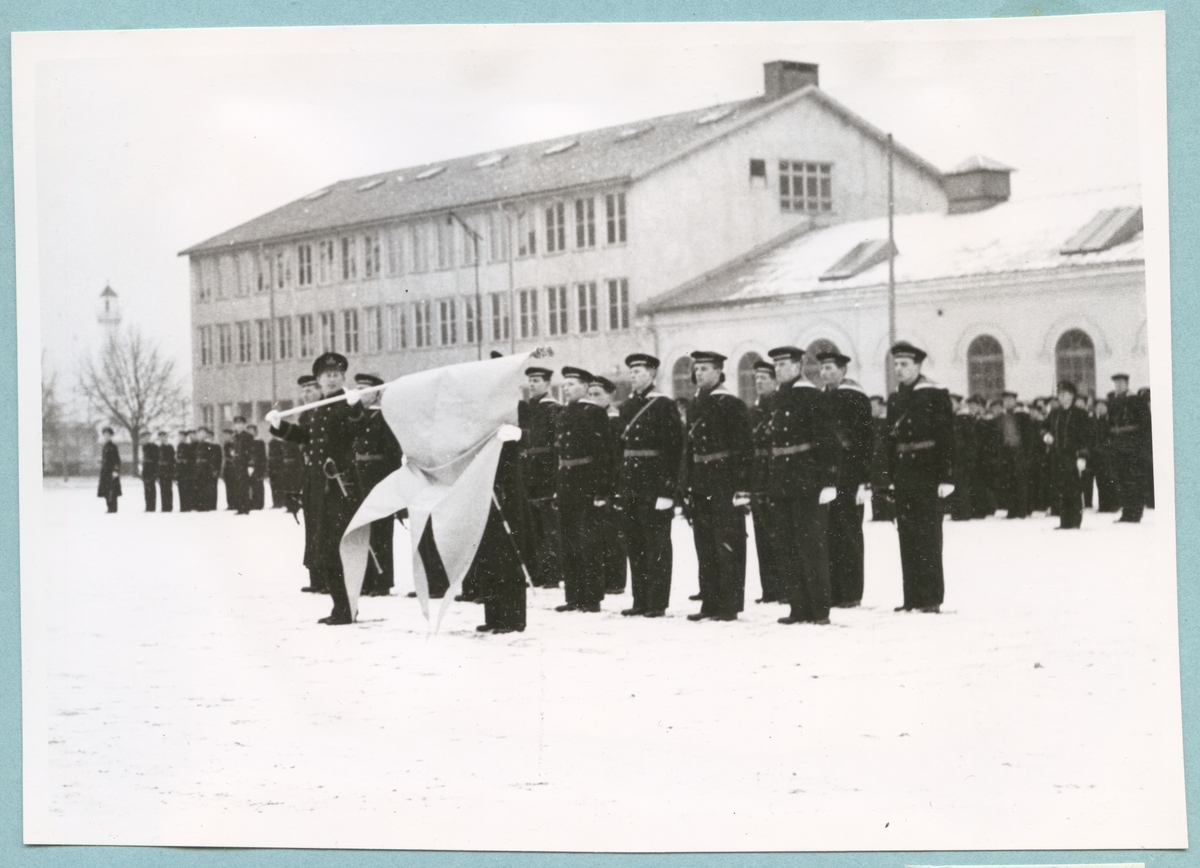 Uniformsklädda flottister står uppställda i snön på bataljon Sparres kaserngård. Längst fram står en militär och en fänrik och håller i en stor flagga. I bakgrunden syns några av bataljonens byggnader.