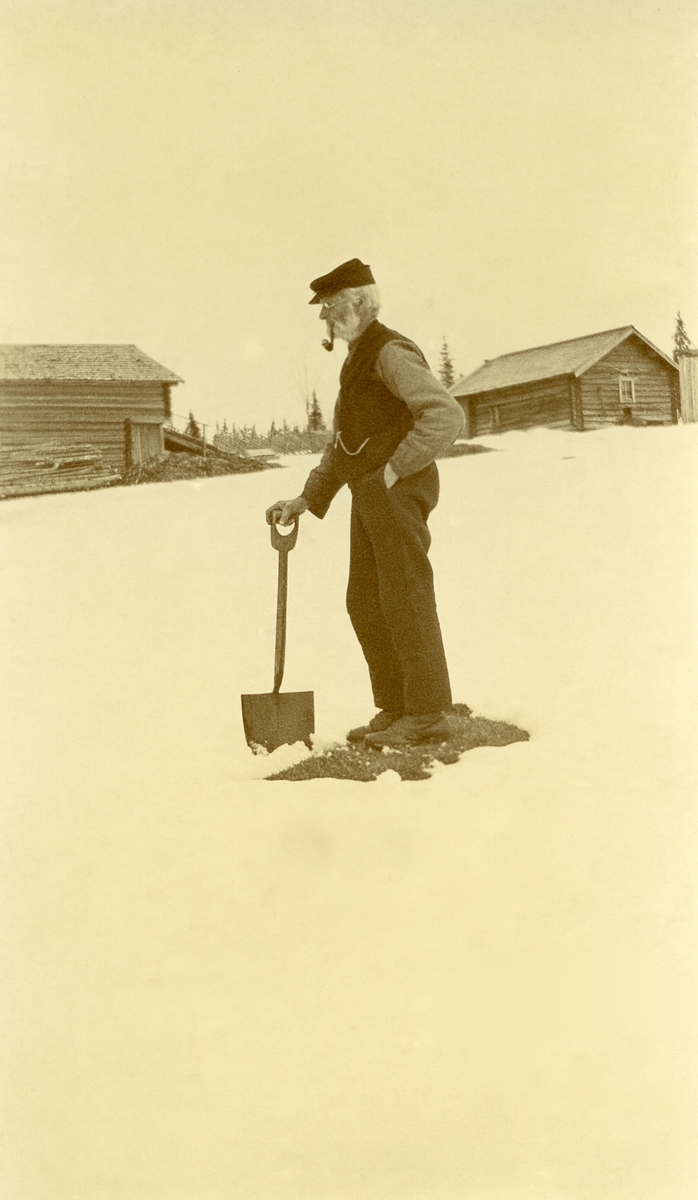 Mann med spade og pipe i munnen stående på bar flekk, midt i snøen, i Langtjønnåsen, Sæter (?) Hus i bakgrunnen.  John Olsen Sæter (Jonstø-Jon), 80 år gammel (1845 - 1930)