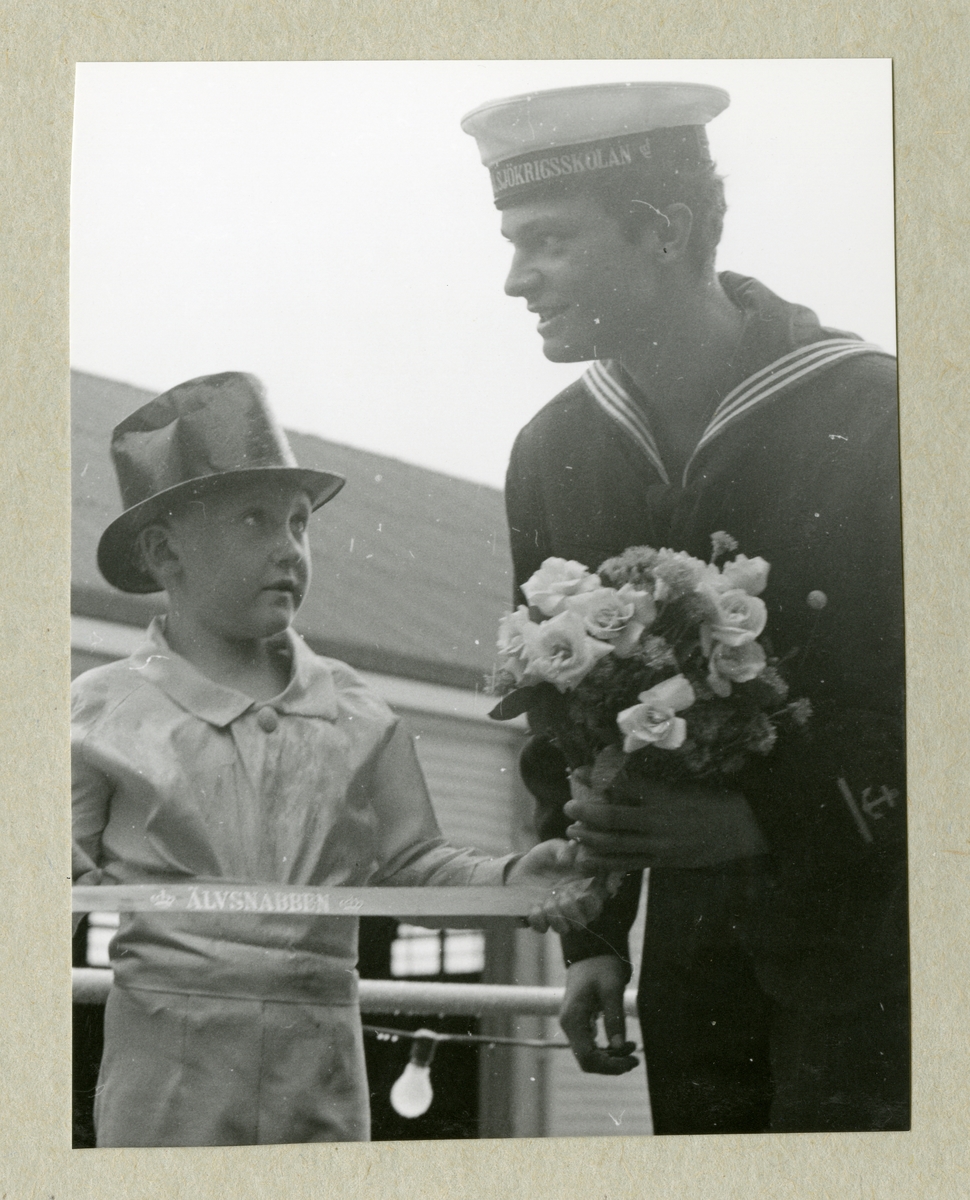 Bilden föreställer en liten pojke i hatt och Carl XVI Gustaf, klädd i uniform. I ena handen håller Carl XVI Gustaf en blombukett. Bilden är tagen under minfartyget Älvsnabbens långresa 1966-1967.