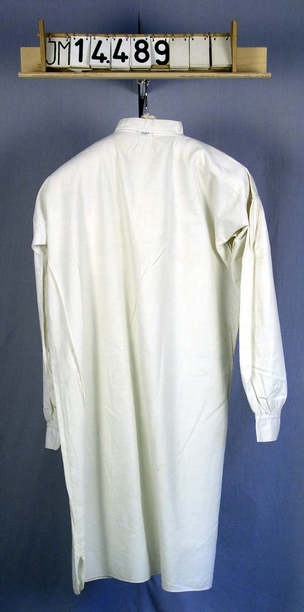 Lång skjorta av ganska vit grov lärft eventuellt av halvlinne, långa ärmar, sprund fram, en metallknapp.