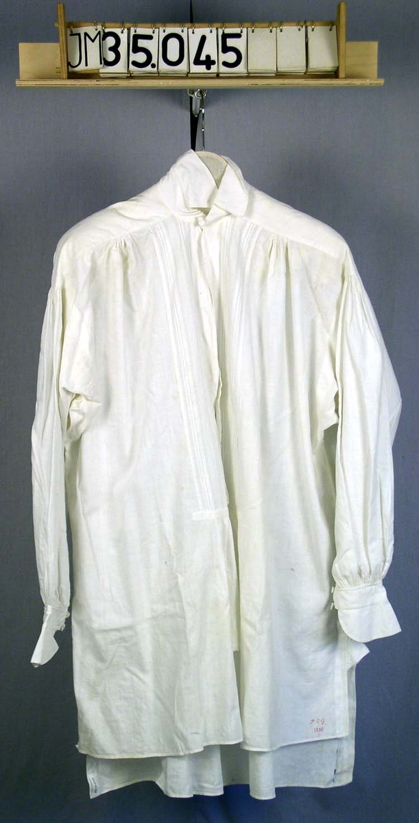 Skjorta av tunnt halvlinne, långa ärmar, skjortbröst med små sydda stråveck, trådknappar. Märkt i nedre högra hörnet fram PCG 1858.