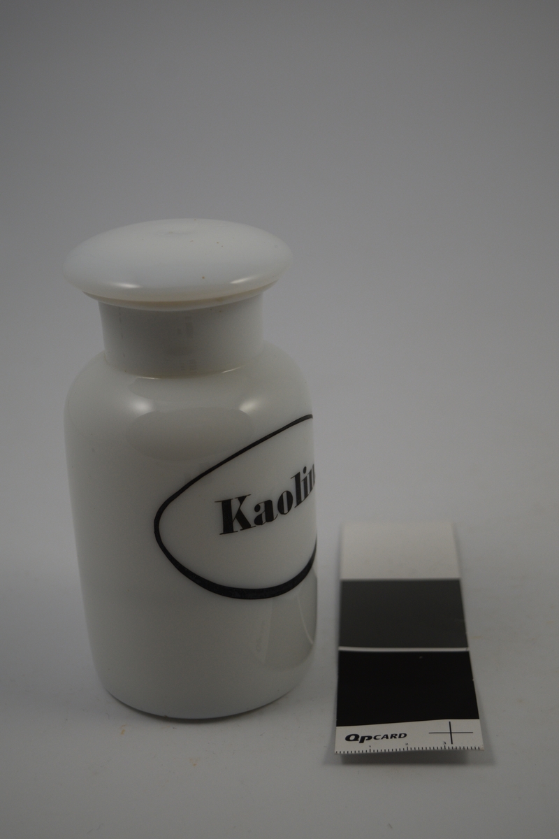 Hvit porselenskrukke med hvit propp. Påsatt etikett med sort oval ramme og sort skrift. Kaolin er syrenøytraliserende og ble brukt mot mageproblemer.