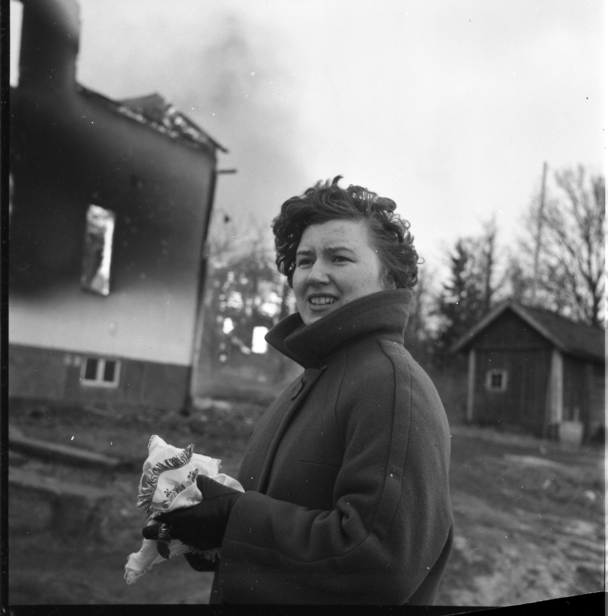 Okänd kvinna med en duk (?) i handen, framför brinnande byggnader i Björkenäs, februari 1964.