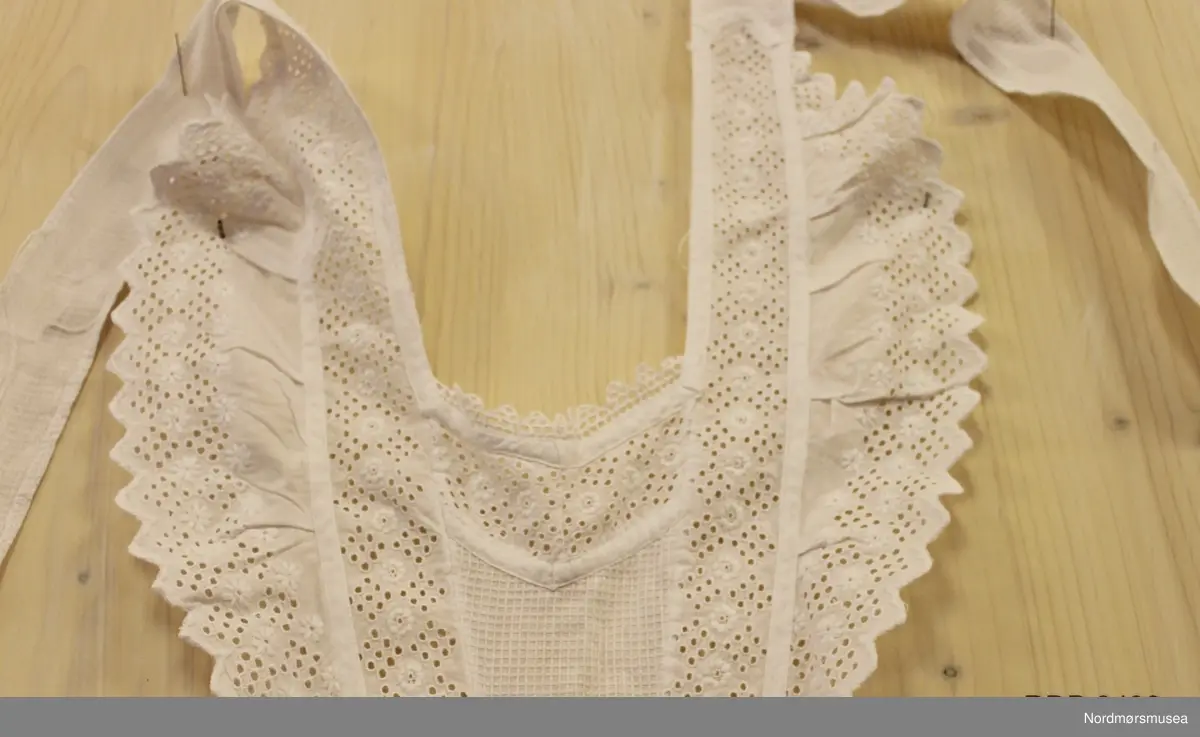 Kvitt serveringsforkle i bomull med innfeldte blonder og blondekant rundt. Smal midje og smekke. Knytteband rundt nakken og midja. Maskinsydd.