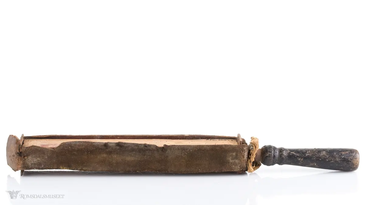 Rektangulær med dreid trehåndtak. Lærrem som går rund hele. Avstivet med metallbeslag foran og bak. Brukt til å stroppe/kvesse barberknivblad før bruk.