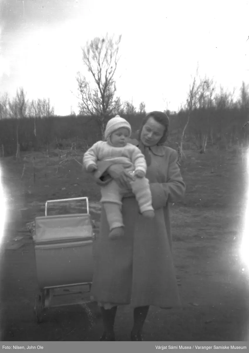 Berit Hansen holder sønnen sin Øystein. Ved siden av henne står barnevognen, trær i bakgrunnen. Fuoitnjárga / Bunes i 1952/53.