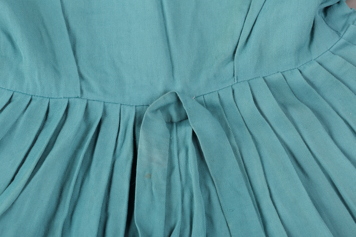 Form: Innsvingt liv, korte ermer, plissert skjørt, fastsydd belte.
Krave.
Bærestykket har vertikale stolper på hver side fra skulder ned til liv, med sydde legg.
Åpning foran med trykknapper. 3 pynteknapper utenpå.