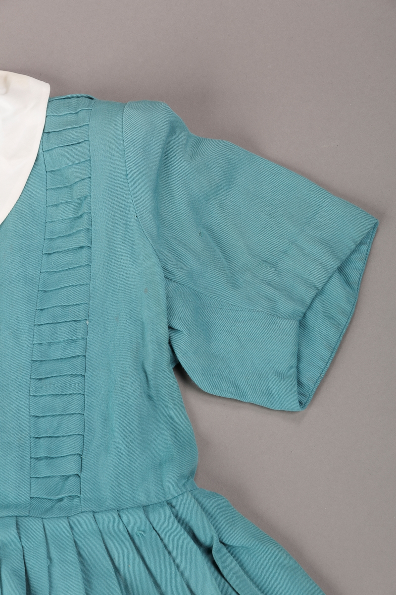 Form: Innsvingt liv, korte ermer, plissert skjørt, fastsydd belte.
Krave.
Bærestykket har vertikale stolper på hver side fra skulder ned til liv, med sydde legg.
Åpning foran med trykknapper. 3 pynteknapper utenpå.