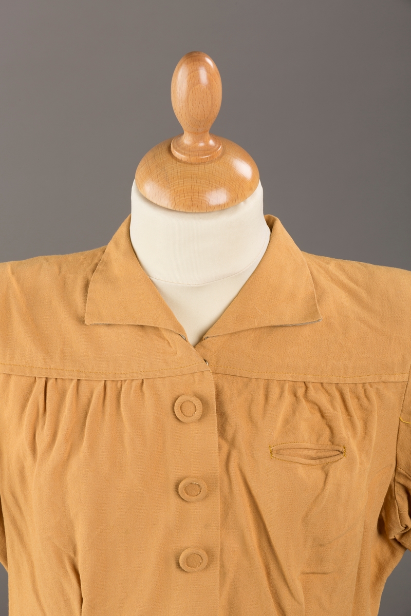 Form: Korte ermer, livet rynket til bærestykke foran, plissert skjørt, tre pynteknapper, en trykknapp, forlomme.