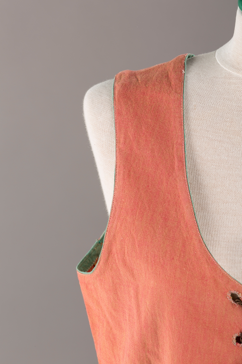 Rustrødt liv med grønn kant og fór, seks skjøter. Håndsydd forsterkning i kontrastfarge øverst i skjøtesplittene. Lukkes med tinnhekte og silkesnor.