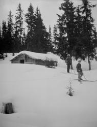 To menn på ski ved tømmerhytte.