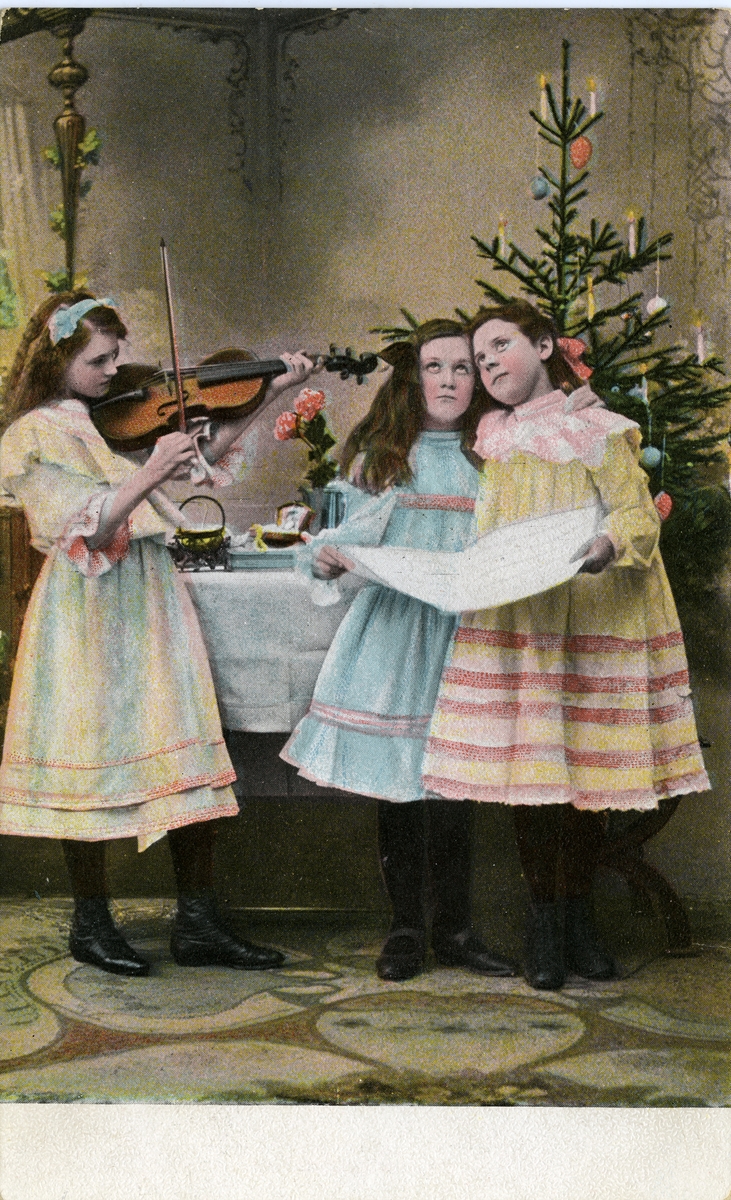 Julekort. Jule- og nyttårshilsen. To jenter synger foran juletreet, akkompagnert av jente med fiolin. Datert julen 1906.