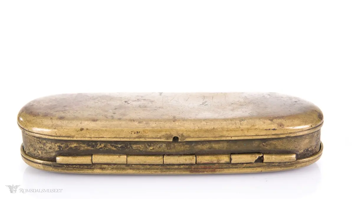 Rektangulær messingeske med avrundede ender. Har en påsatt og påklinket utenpåliggende bunn og utenpåliggende, hengslet lokk