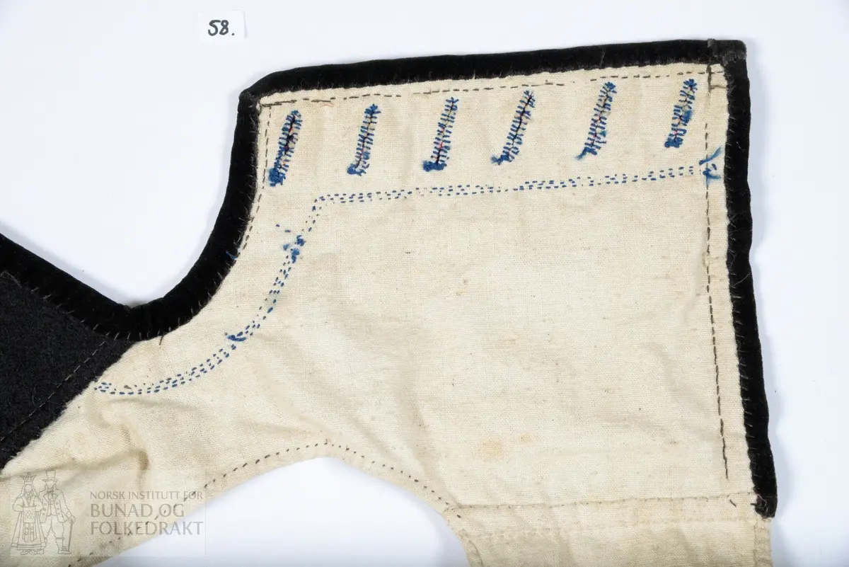 Vest, sauma av silkedroguet. Blå satengbotn med rosa flotterande trådar og innvove små blomster i ulike fargar. Framkant, krage og nedkant er påsydd ein smal svart kant av bomullsfløyel. Innanfor denne i framkant og krage er det påsydd eit breitt stykkje svart tynn fast klede. Denne er klipt med taggar etter sårkanten, pryda med tre tette konturstingsrader med blå ulltråd. 
Kragen er fora med tjukk brunsvart vadmål, sydd med to langsgåande avstivande stikningar. Ryggen er ufora, sauma av litt grov bomullslerret. 
Same type for i framstykkja. 
I ermholet på framstykkjet er foret bretta dobbelt rundt på framsida, og sydd ned med små javne faldesting. Sidesaumane er drage eit godt stykkje inn på kvart framstykkje. 
Eine framkanten har påsydd 6 stk skråstilte knapphol sauma av same type blå ulltråd. Merkjer etter knappefeste gjennom fløyelskanten på andre framstykkjet. 
I sidesaumen er hovudstoff framstykkje og ryggfor sydd saman med attersting, deretter er for framstykkjet bretta inn og sydd ned 1 cm bak den fyrste saumen, med faldesting som syner som prikk på retta.