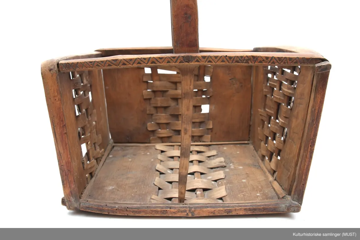 Firkantet trekurv med flettedekor og treskjæring på både kurv og håndtak. Dekoren består av to seksbladete blomster på hver side samt ulike trekantmønstre langs kanten av kurven og håndtaket. En av sidene i kurven mangler.