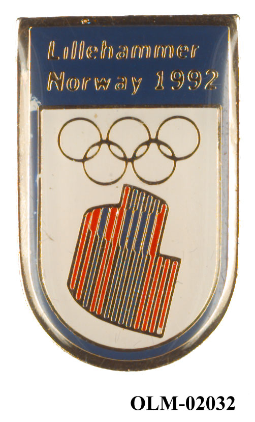 Skjoldformet merke med de olympiske ringer og nordlyslogo som vender nedover på hvit bakgrunn. Tekst øverst i sølvbokstaver på mørkeblå bakgrunn. Merket har gulnet.