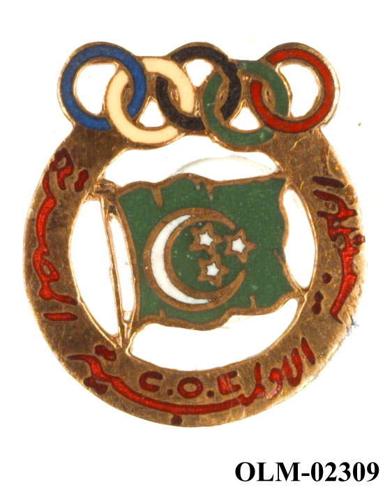 Rundt merke med de olympiske ringer øverst. I midten det et grønt flagg med halvmåne og tre stjerner. Arabisk tekst på sirkel rundt flagget..
