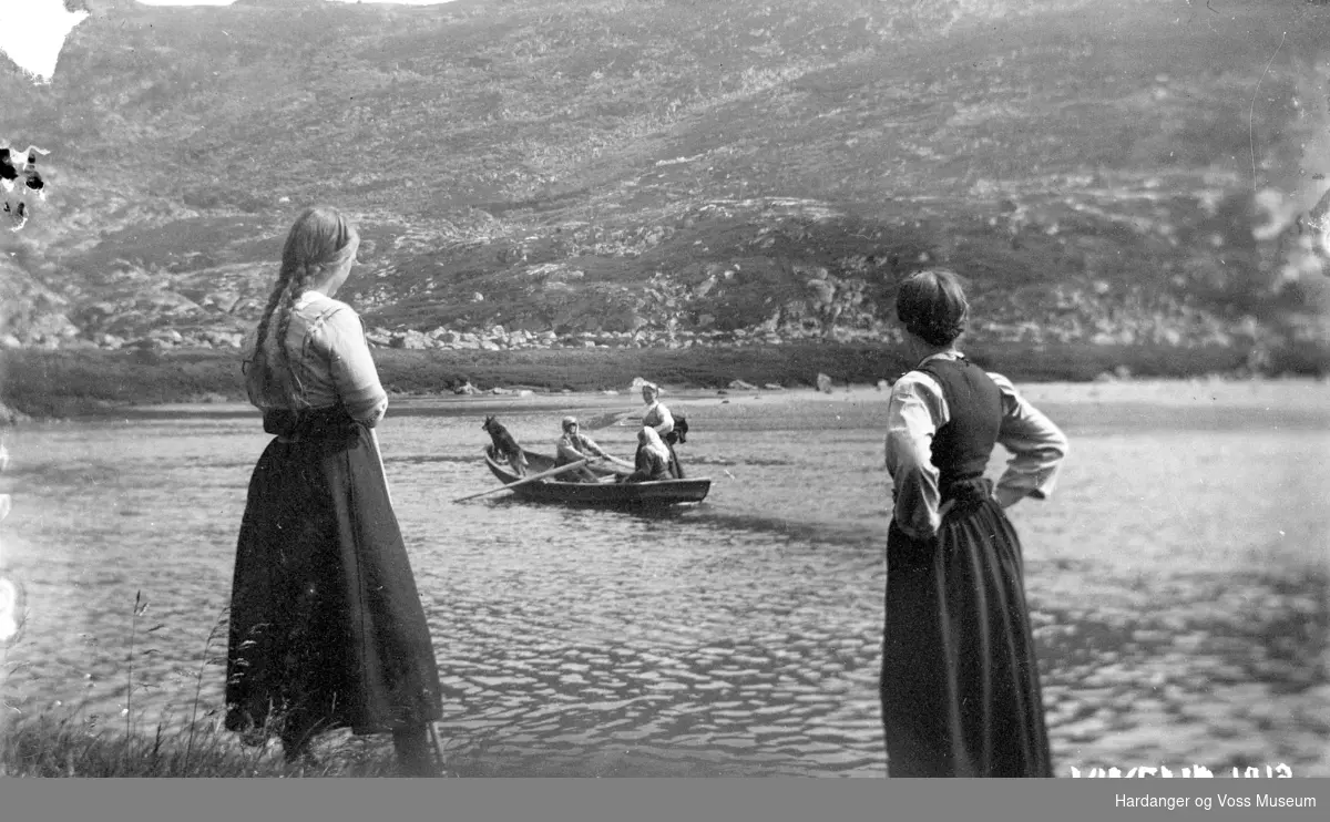To kvinner ser på ein robåt med tre personar i ro over elva