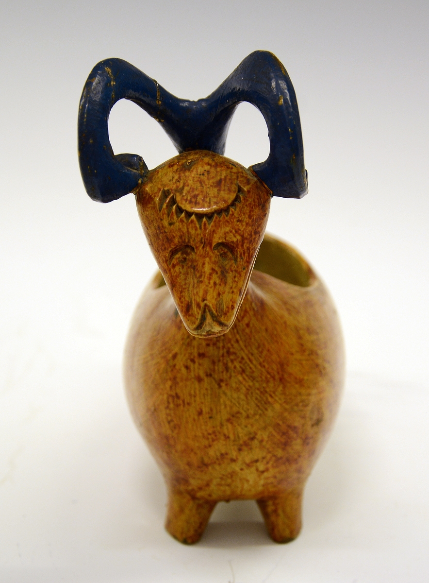 Kane. Fra protokollen: Kane, saak. "ølbøkaar", av træ. Kroppen nærmest mindende om gris + and, med 4 avstumpede ben og avstumpet hale. Hovedet bærer 2 foroverbøiede, fritliggende væderhorn (værhorn), som med sin forreste krumning hviler paa kjæven; hovedet forøvrigt væderlignende. Sen maling, kirketræsfarvet krop, blaa horn, hvit inside.