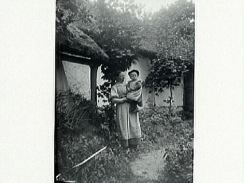 En kvinna med ett barn på armen står vid ett ålderdomligt hus med halmtak. Barnet bär basker. Längs husväggen står två späda lönnar i vildvuxen grönska. 1890-tal.
