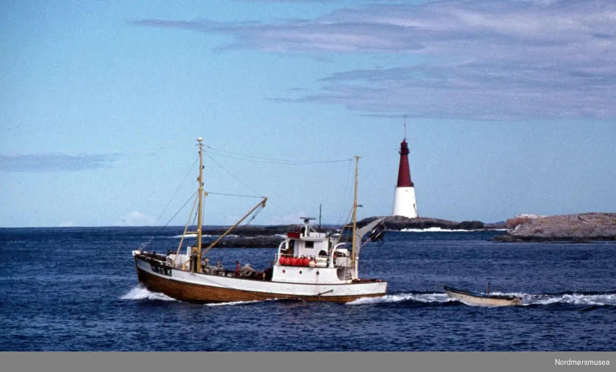 Foto av en fiskebåt med fyrtårnet like utenfor fiskeværet Grip i Kristiansund kommune. Fotograf er Kjell Haug og er den som står bak samlingen med fargefoto/dias fra Kristiansund og Grip. Bildene dateres til omkring 1950-1970. Fra Nordmøre museums fotosamlinger.