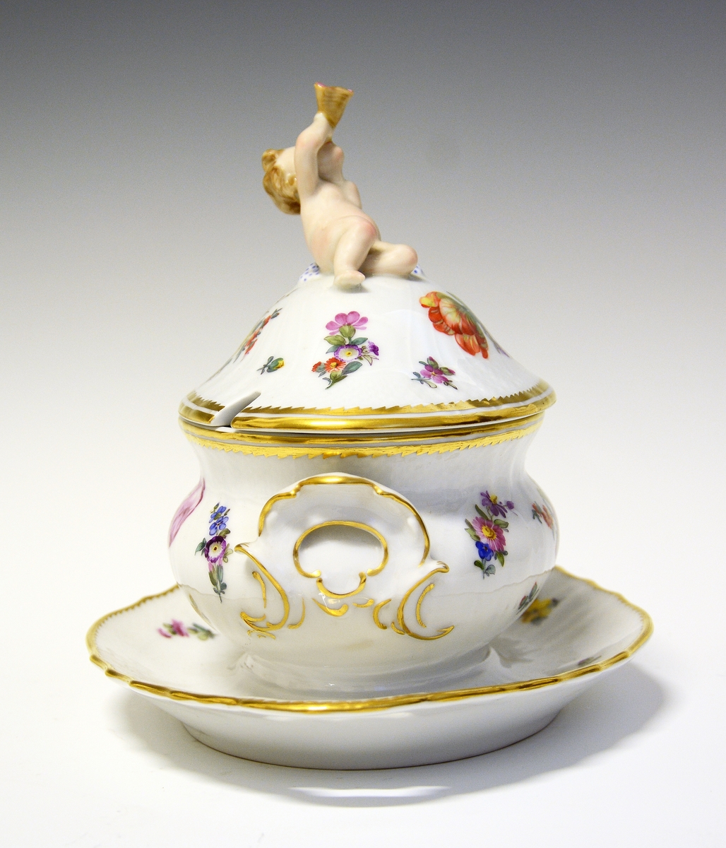 Sauseterrin av porselen dekrorert med håndmalt blomsterdekor og modellert figur av et barn med blåseintstrument som lokknapp. Hanker på hver siden med skjellform. Alle kanter er markert med gull.