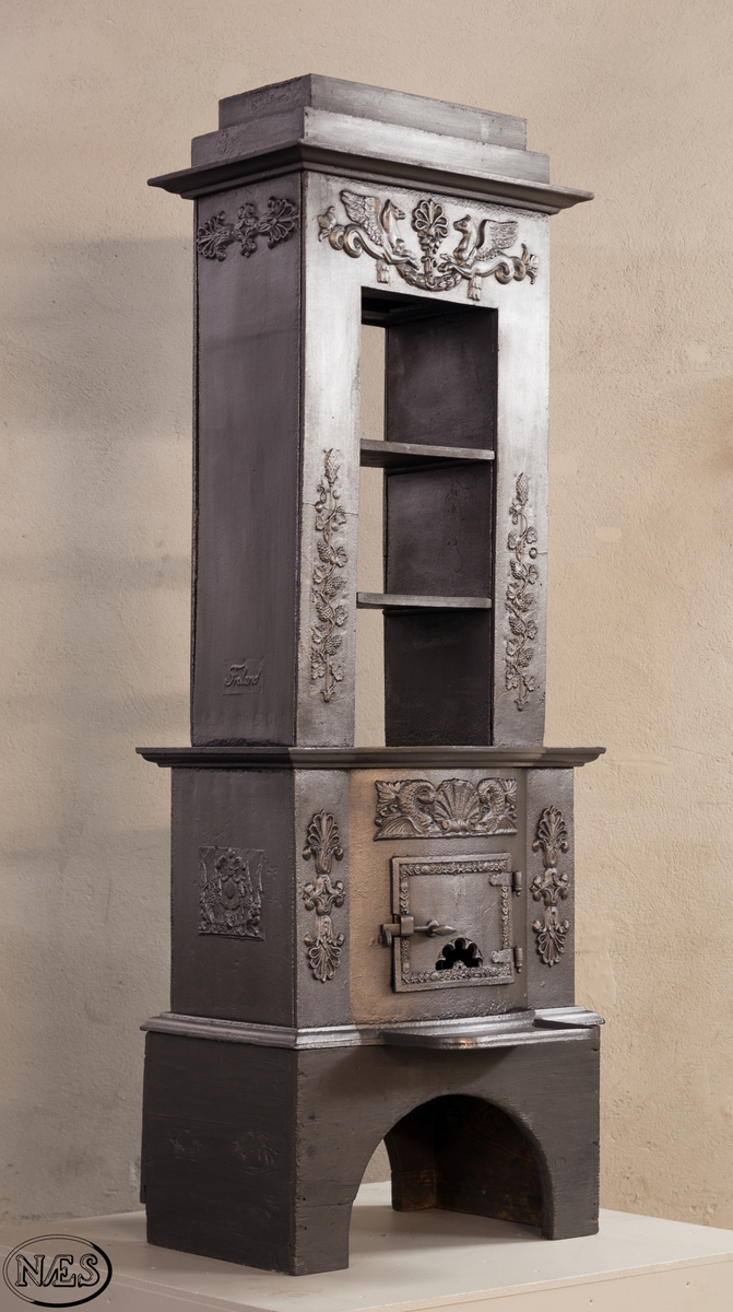 Ovnen består av to avdelinger. Underdelen har et frembuet midtparti med kamskjell flankert av sjødyr på hver side. På kortsidene skjold, løvehode og symmetriske ørner. På bakplate en krukke med symmetriske engler.
Overdelen er høy, firkantet med tredelt åpning. Empire ornamentikk og avtrappet topp. Øverst på frontplaten symetriske fabeldyr (pegasus) vendt mot hverandre.