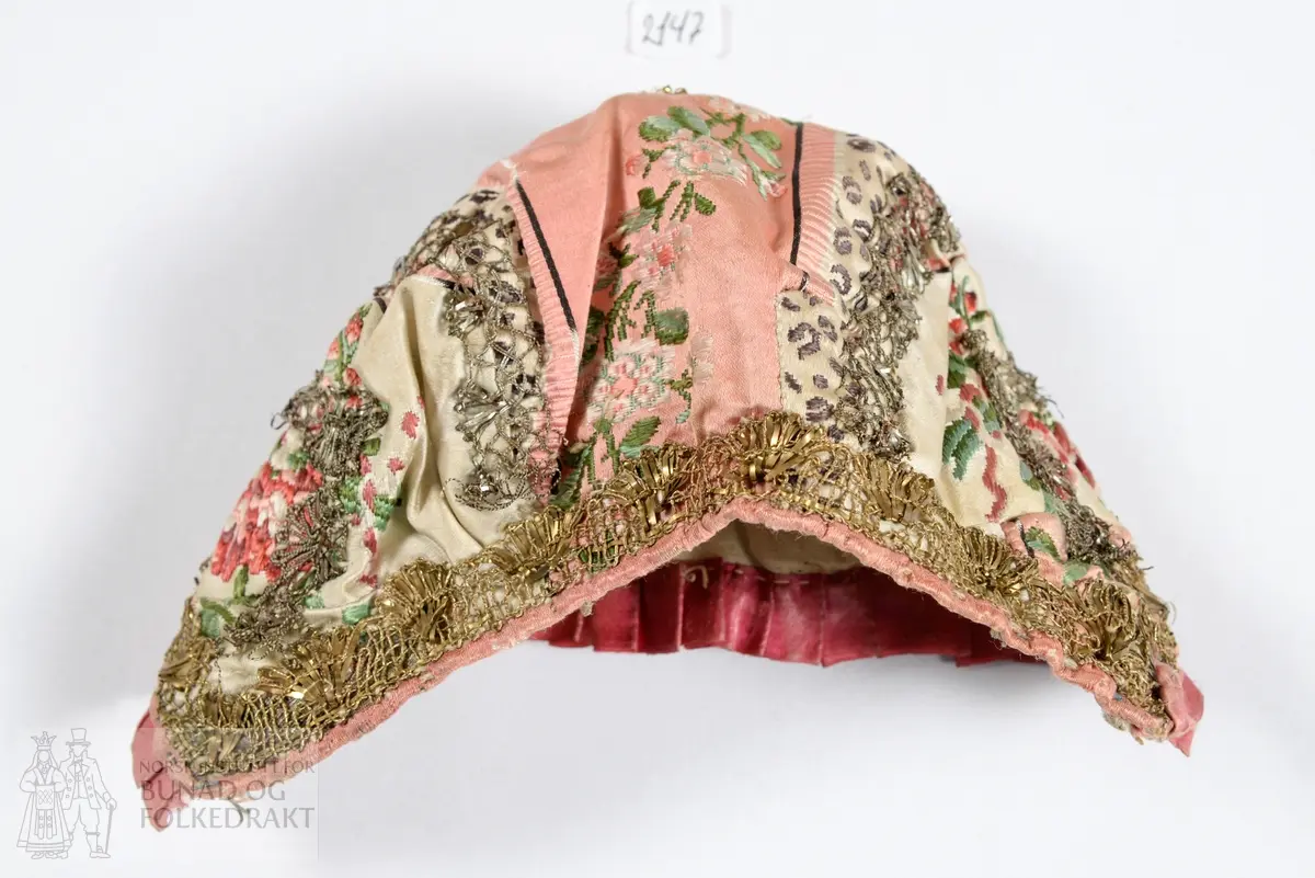 Dåpslue. Av rosa og hvit silkedamask med brosjert blomstermønster i to ulike rosa nyanser og grønt. Dekorert med metallkniplinger, i både sølv og gull. Smal rosa silkekanting rundt hele lua, i tillegg er et mørkerosa silkebånd i satengbinding foldelagt langs framkanten.  Foret med naturfarget linlerret. For og lue er vrangsydd, og så sydd sammen. Sømmene fra kniplingene er synlige på vrangen. Kun håndsøm. Stoffet er utnyttet med flere små skjøter.