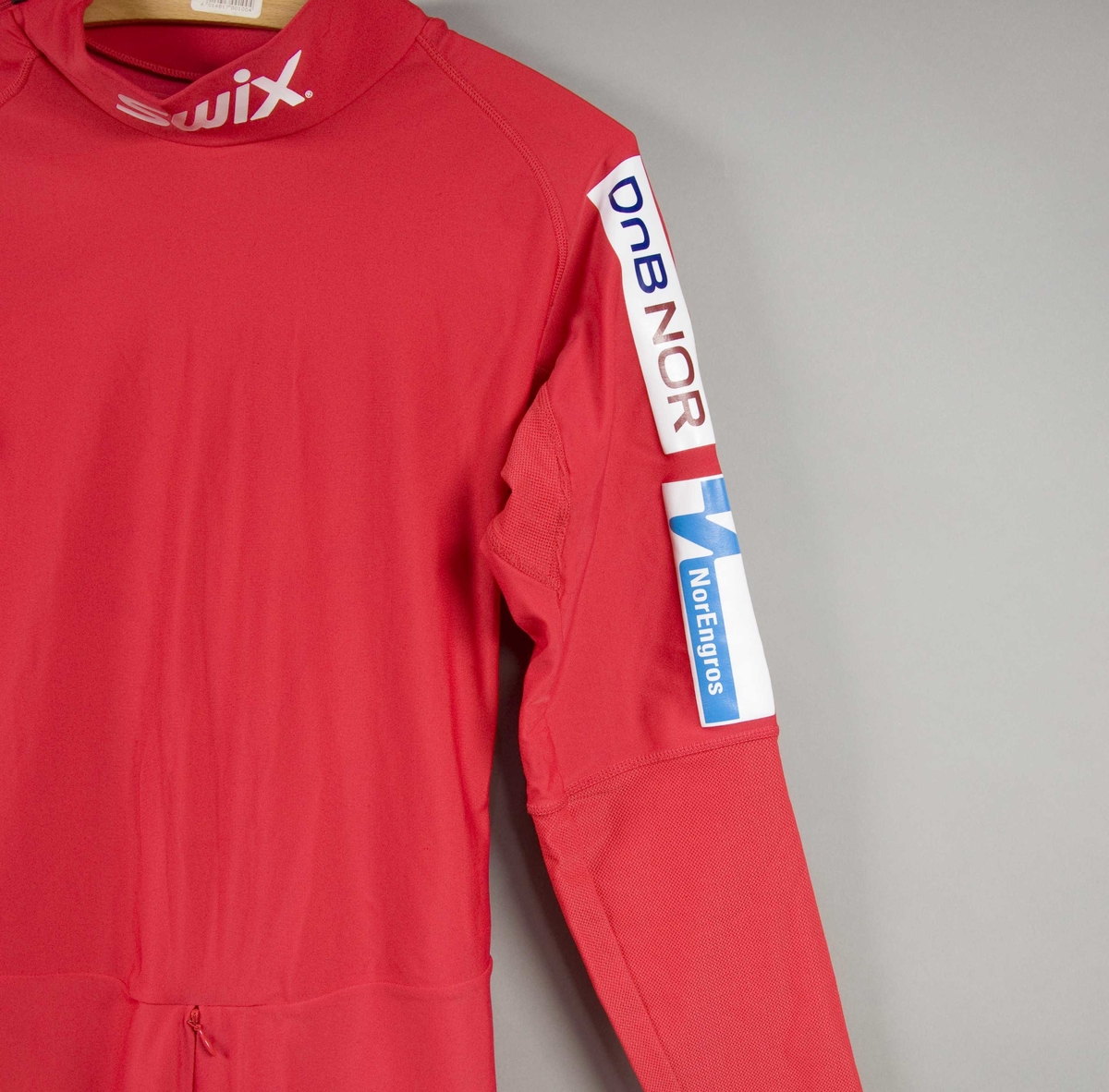 Rød langrennsdress med hvite og blåe striper. På dressen er det flere laminerte logoer for forskjellige sponsorer.