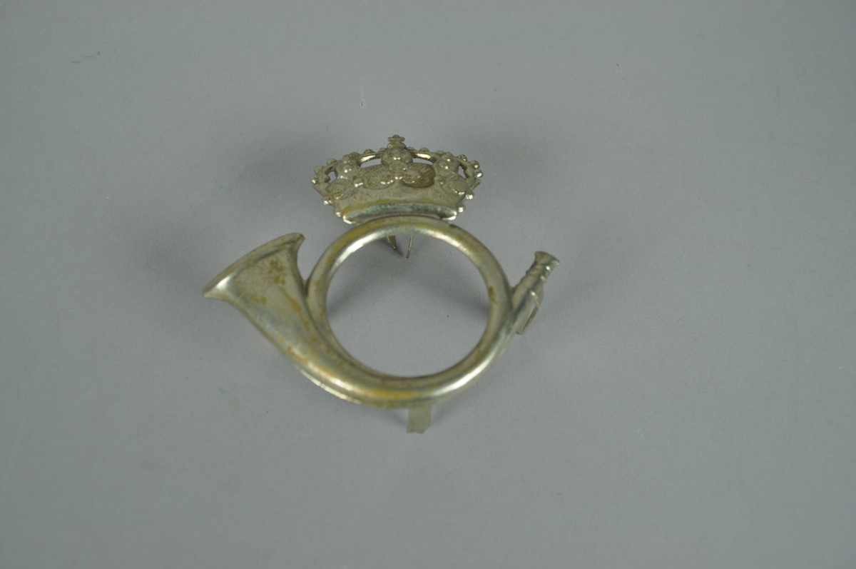 Posthornmerke i sølvfarget metall.