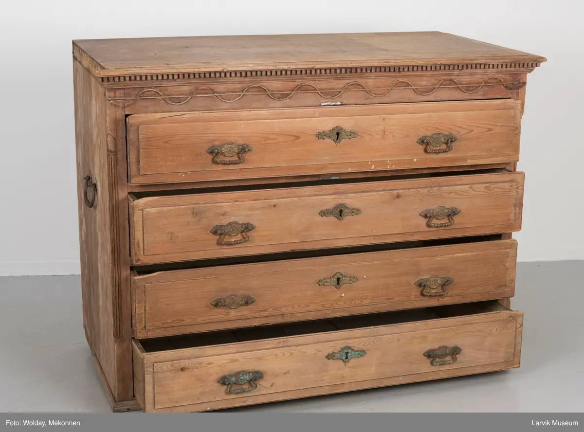 Kommoden har 4 skuffer. 2 beslag for å dra ut, 1 beslag for nøkkelhull, messing. Utskåret dekor under øvre pyntelist "tanngard" rundt på 3 kanter. Øvre skuff utskåret med bølgebånd med rett linje som bryter bølgene. Utskjæring kun i front. To av kommodens hjørner har utskåret søyle. Håndtak på kortsidene for å bære i.