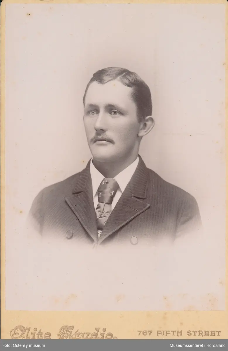 portrettfotografi av ung mann med bart, stripa jakke, kvit skjorte og mønsta silkeslips