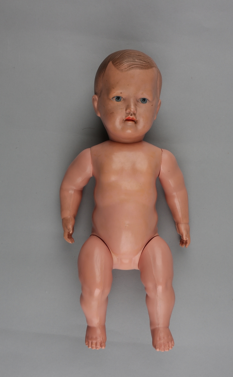 Babydukke i plastmateriale. Armer og ben festet med elastisk tau. Påmalt hår, øyne og munn.