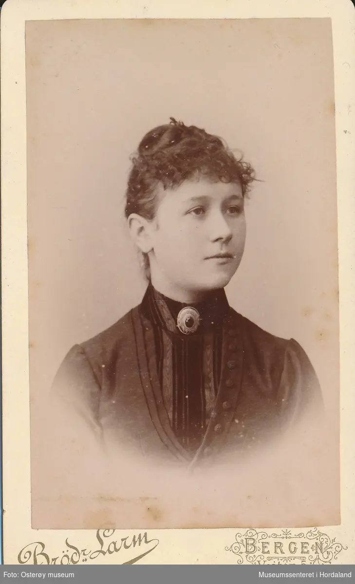 portrettfotografi av ung kvinne/jente med hårtopp, krølla lugg, kledd i einsfarga jakke med plissert innfelling og brosje i halsen