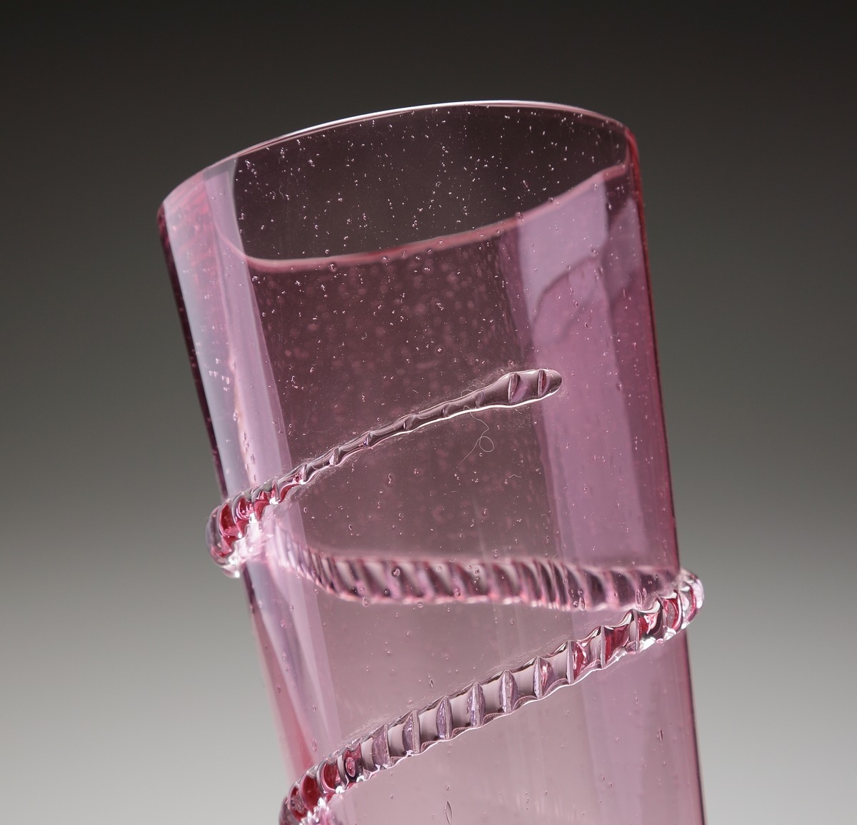 Vas. eller möjligen pokal.
Stilen på glasen är gjord så att den ska se ålderdomlig ut.
Hög rosa, konisk vas med spunnen och naggad ofärgad glastråd. 
Foten rosa  och konisk med markerade ränder.