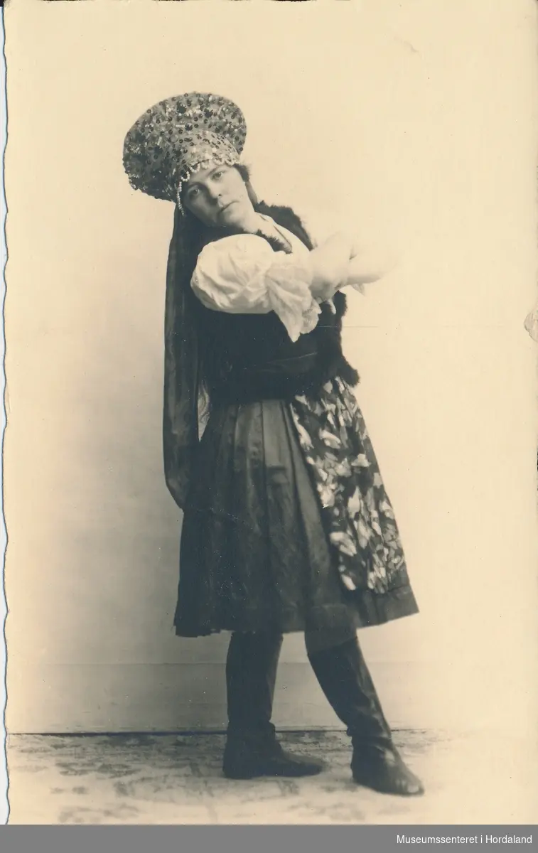 atelierfotografi av ung kvinne i folkedrakt med stor rund krune med slør, kvit bluse, mørk vest og skjørt, blomstra forkle og høge støvlar