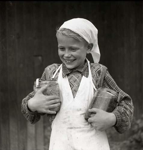 Pojke i kökskläder bär två glasburkar med inläggningar. Unga odlare 1948 - 1949.