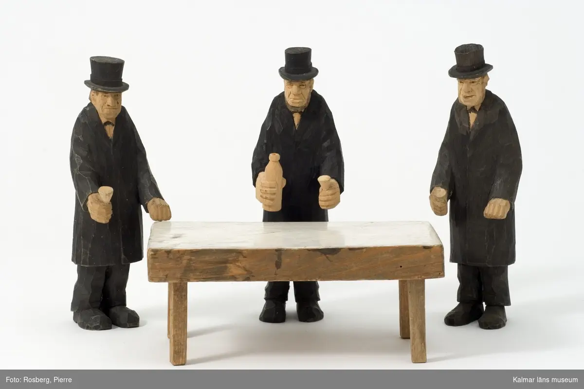 KLM 43830:12 Skulptur, av trä. Benämnd, Gravöl. Datering, ca 1930. Signatur, HC. Figuren består av tre svartklädda män iklädda hög hatt, de står vid ett bord med målad vit duk och har varsin öl i handen.