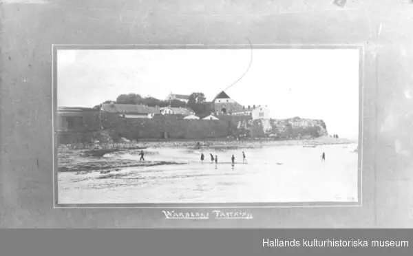 Fotografi. Varbergs fästning från nordväst. I förgrunden lekande barn i vattnet, "Barnens badstrand". Färgretuscherat.