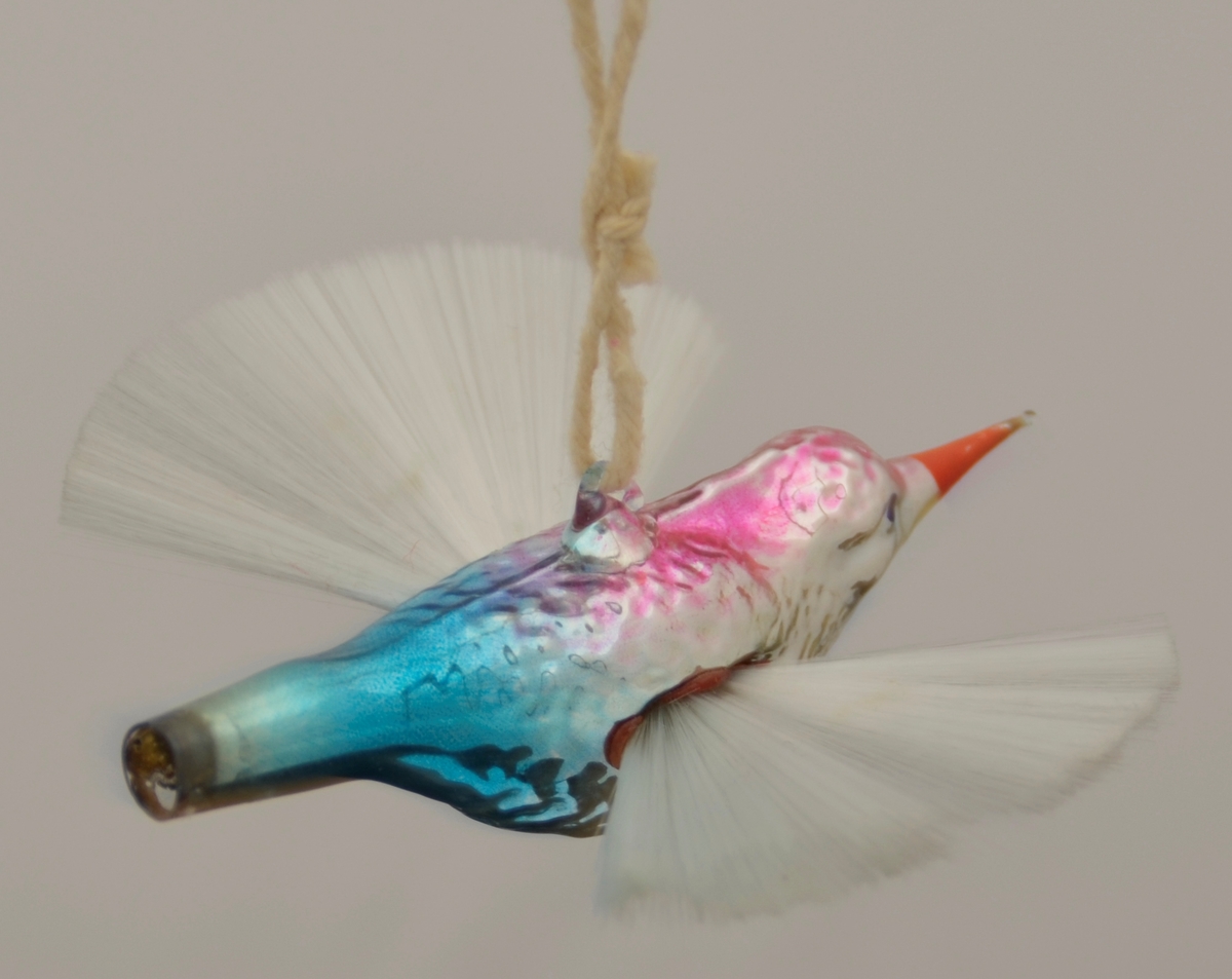Juletrepynt. Glasfugl med vinger som ei vifte av silkeaktig, stiv tråd. Glaset er sølvfarga i botn med lilla og blått på kroppen og raudt nebb. Tråd gjennom oppheng på ryggen.