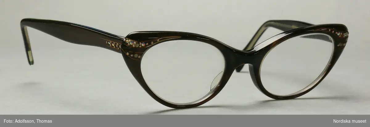 Ett par glasögon i brun och svart plast med mineralglaslinser (H+0.5, V+0.5). Glasögonen är bruna på framsidan samt på utsidan av skalmarna och svarta på insidan av bågen och skalmarna. Strassdekor på fronten vid gångjärnen samt på skalmarna vid gångjärnen.  På insidan av skalmarna står det instansat "REVELLEE  U.S.A.  5 1/2". På bågens insida står det instansat "44-20".
/Petrine Knight, 2014-12-03