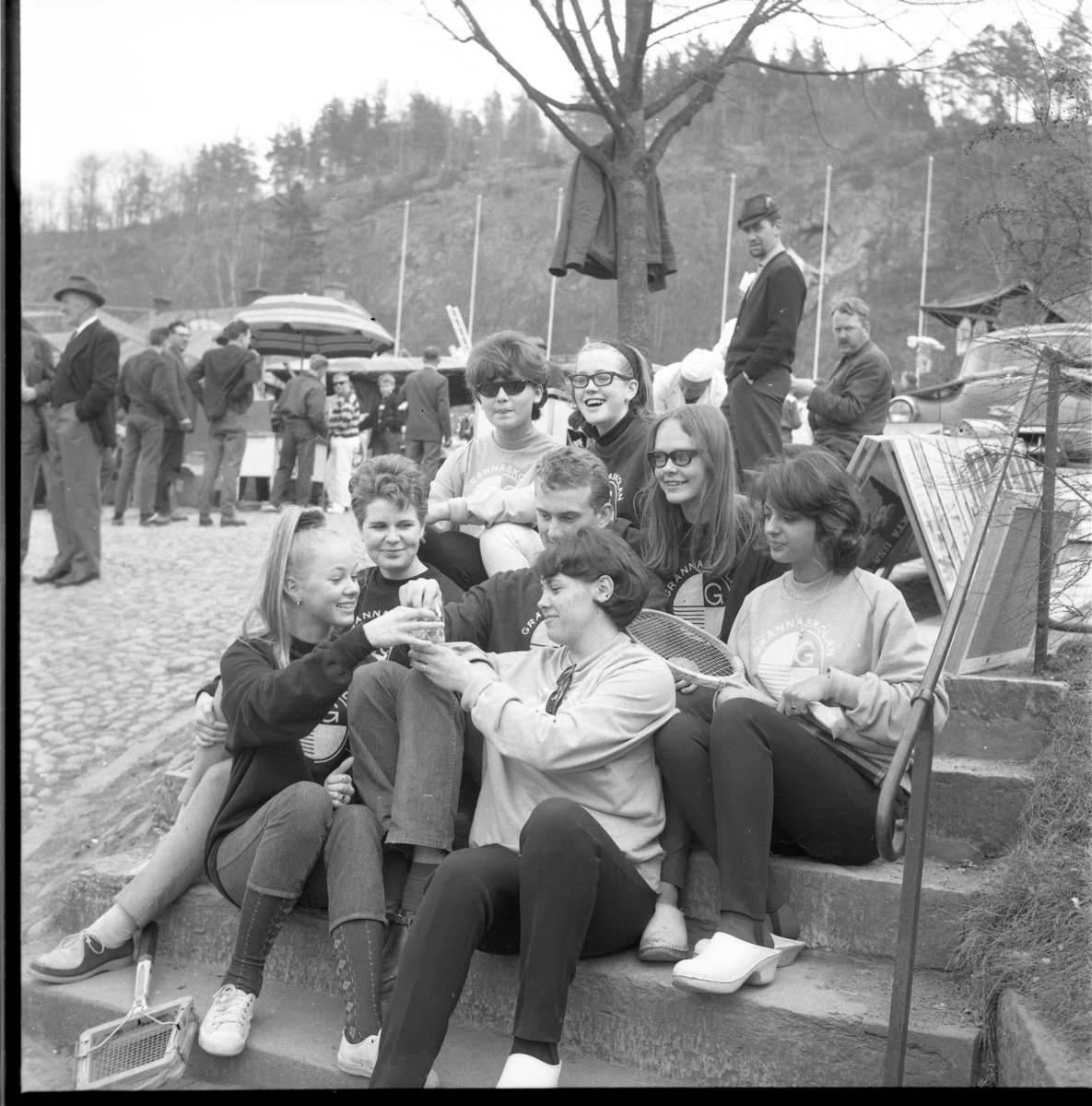 Grännaskoleelever södra trappan vid Gränna torg, 1964. Längst ned till vänster sitter Marita Wallmo, högst upp till höger sitter Monica Granbom och snett nedanför henne med glasögon sitter Lena Blomqvist. De äter godis ur en påse. En flicka har ett tennisrack i knät. 
Det är marknad på torget
