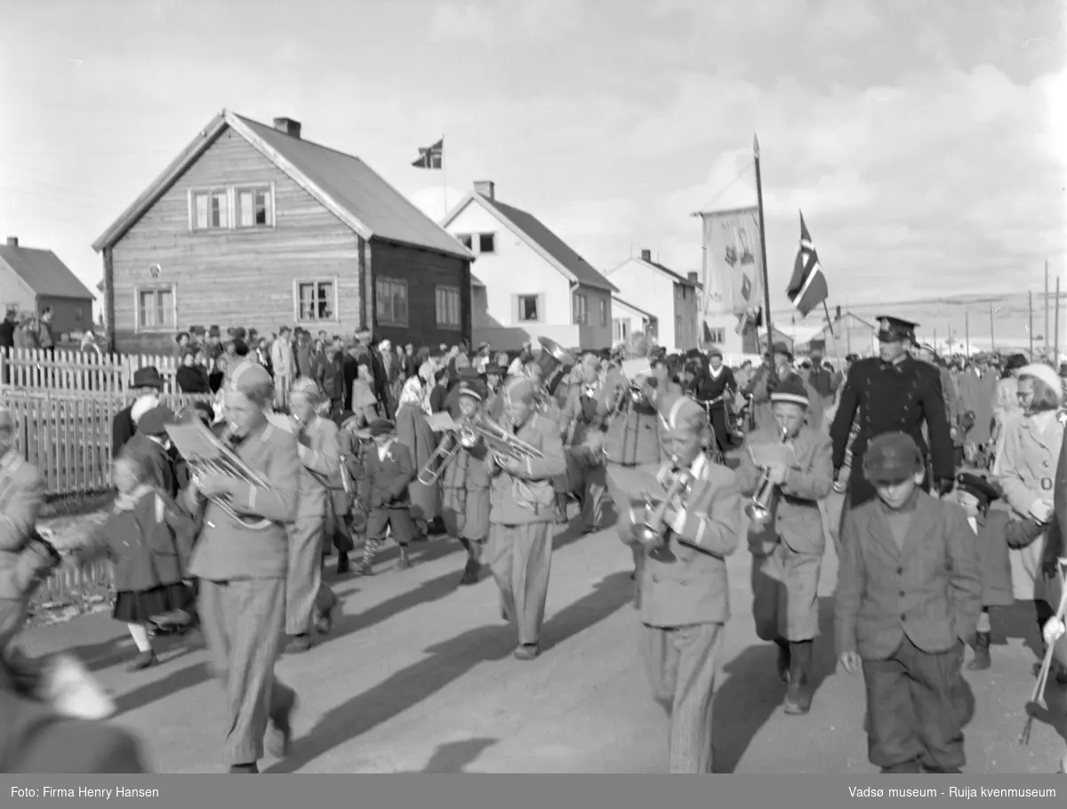 Vadsø 17 mai 1952. 17.mai-toget går gjennom Oscarsgate fra øst mot vest. Ser fjellet Melkevarden i bakgrunnen. Guttemusikk-korps med uniformer spiller. Politimann, faner og flagg.
