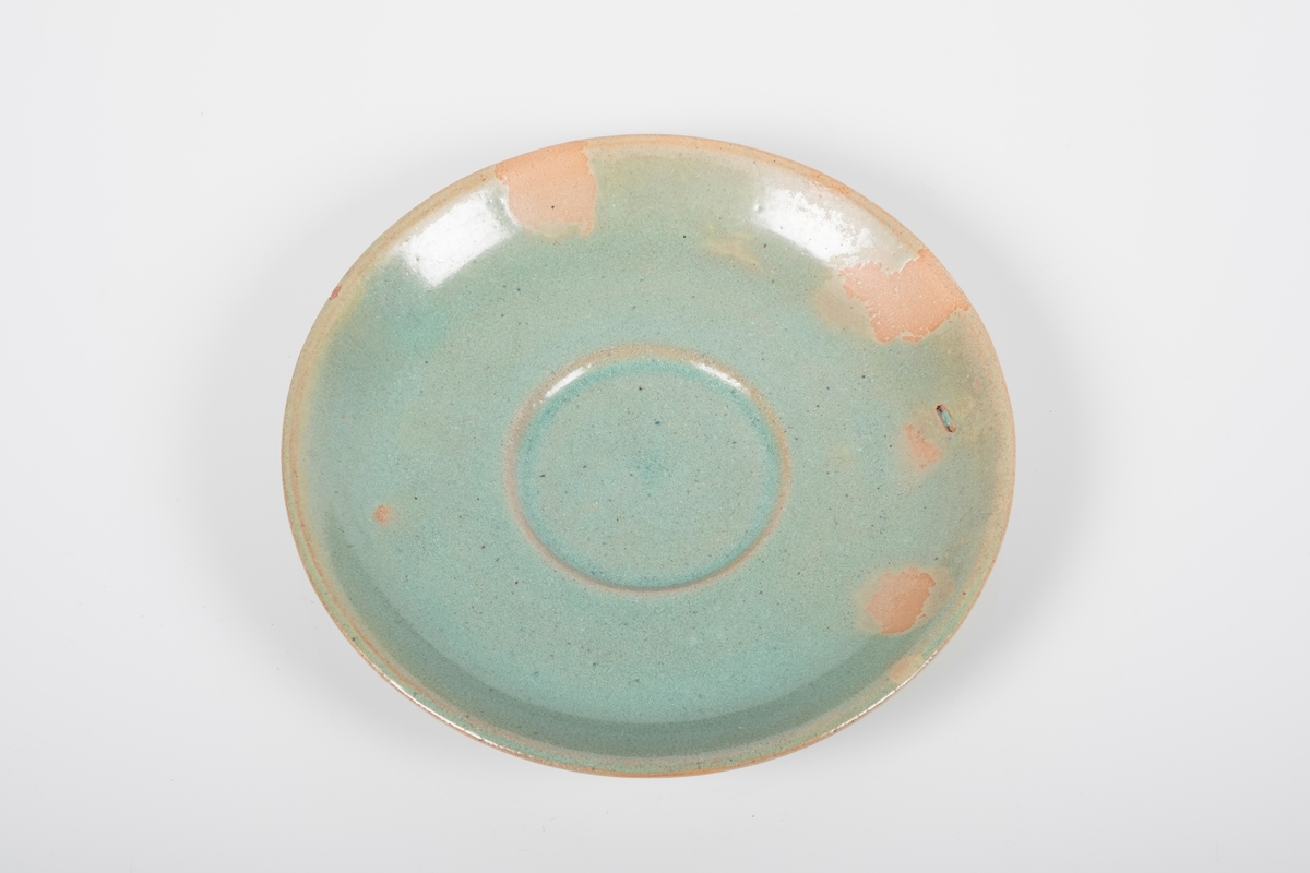 Rund skål i keramikk med grønn lasur. Skålen har spor etter tre knotter på bunnen, usikker funksjon. En hakk på oversiden (produksjonsfeil).