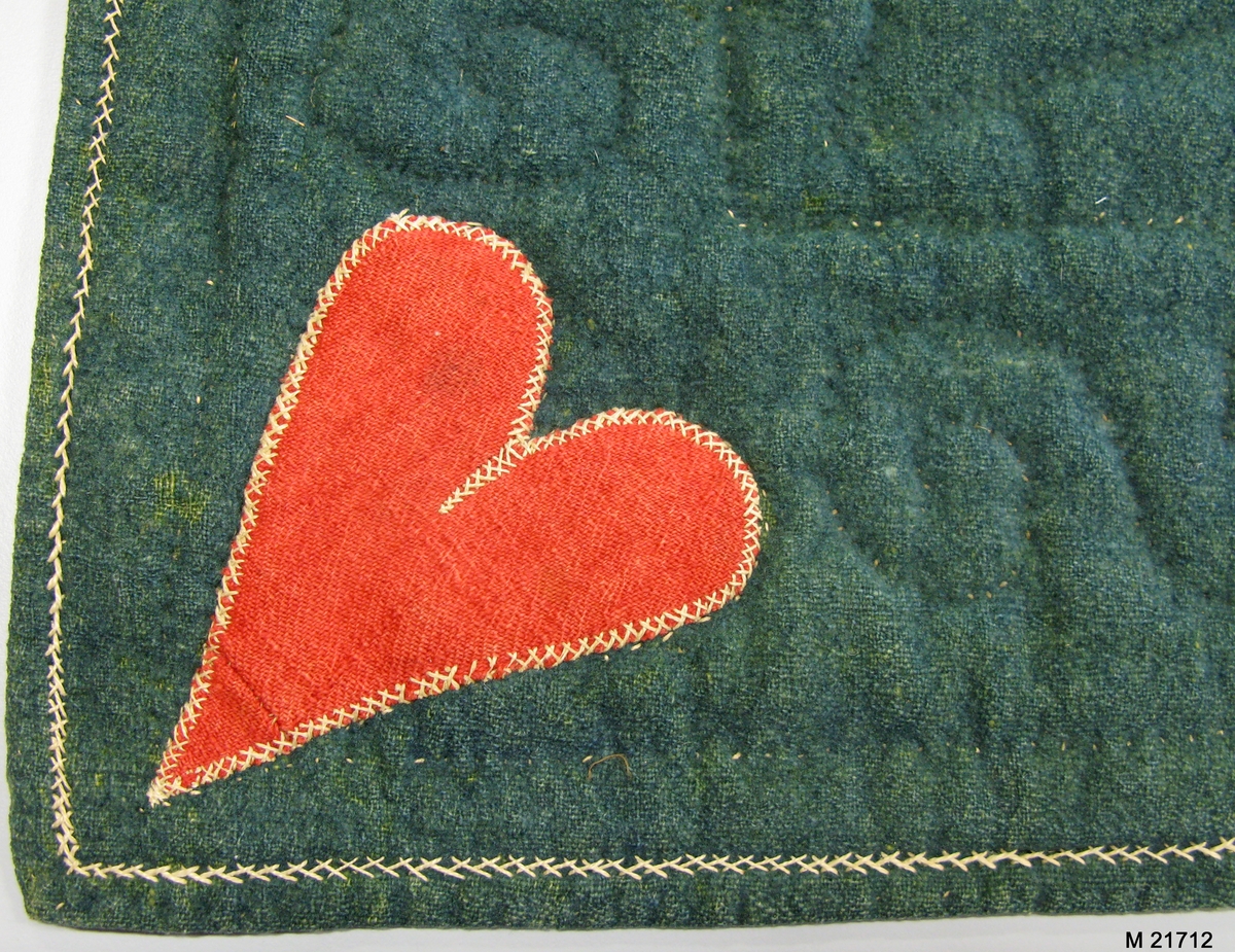 Täcke av grön 2-skaftad och röd 3-skaftad vadmal. Grön bred bård med röda hjärtan i påläggsöm (applikationsbroderi), i mitt mindre röd spegel med gröna påläggsömmar samt I I D 1818 i flät - och stjälksöm. 
Flätsöm runt täckets kant och vid påläggsömmens konturer. 
Baksida: lärft av vänsterspunnet lingarn, 14 varp - och 10 inslagstrådar i cm. Mellaninlägg av ospunna linblånor, ett glest mönster i sticksöm över hela täcket. All sömnad utförd med högertvinnat lingarn.
Inskrivet i huvudkatalogen 1971.
Var med i Textilinveneringen 1928-30 och är därför även märkt N.3.51 (Primus-id: TIKN3051).